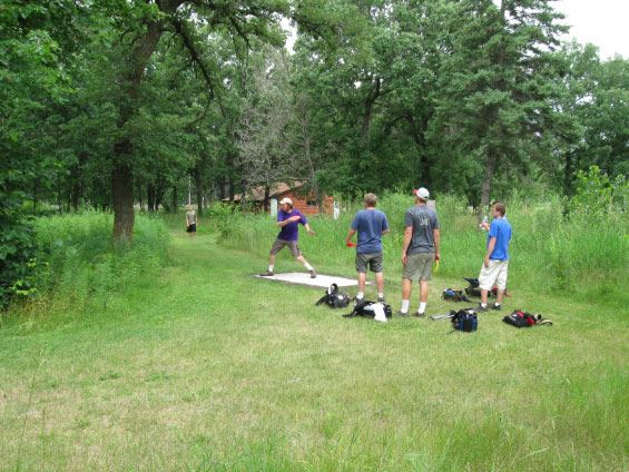 Four men at disc golf stand one throwing